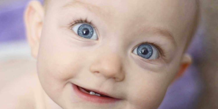 A close up of a baby girl's face as she smiles at the camera. She is 10 months old. She has two bottom front teeth and big blue eyes. A light purple towel behind her is out of focus due to the shallow depth of field. She has bare skin because it is bath time.